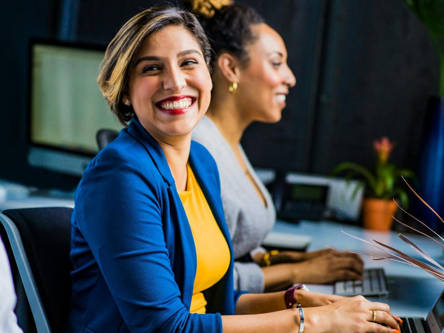 Three diverse professionals working and smiling at office desks, fostering teamwork and collaboration.